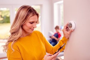 Mother At Home With Son Adjusting Smart Central Heating Thermostat Control With App On Mobile Phone
